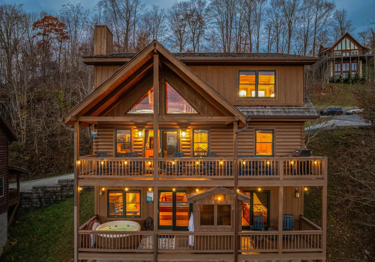 The image captures the exterior of a log cabin at dusk, with a warmly lit balcony adorned with string lights, a bubbling hot tub, and large windows that reveal inviting indoor light. The backdrop features sweeping mountain views, and an outdoor fire pit is visible, suggesting a cozy communal area for gatherings under the sky.