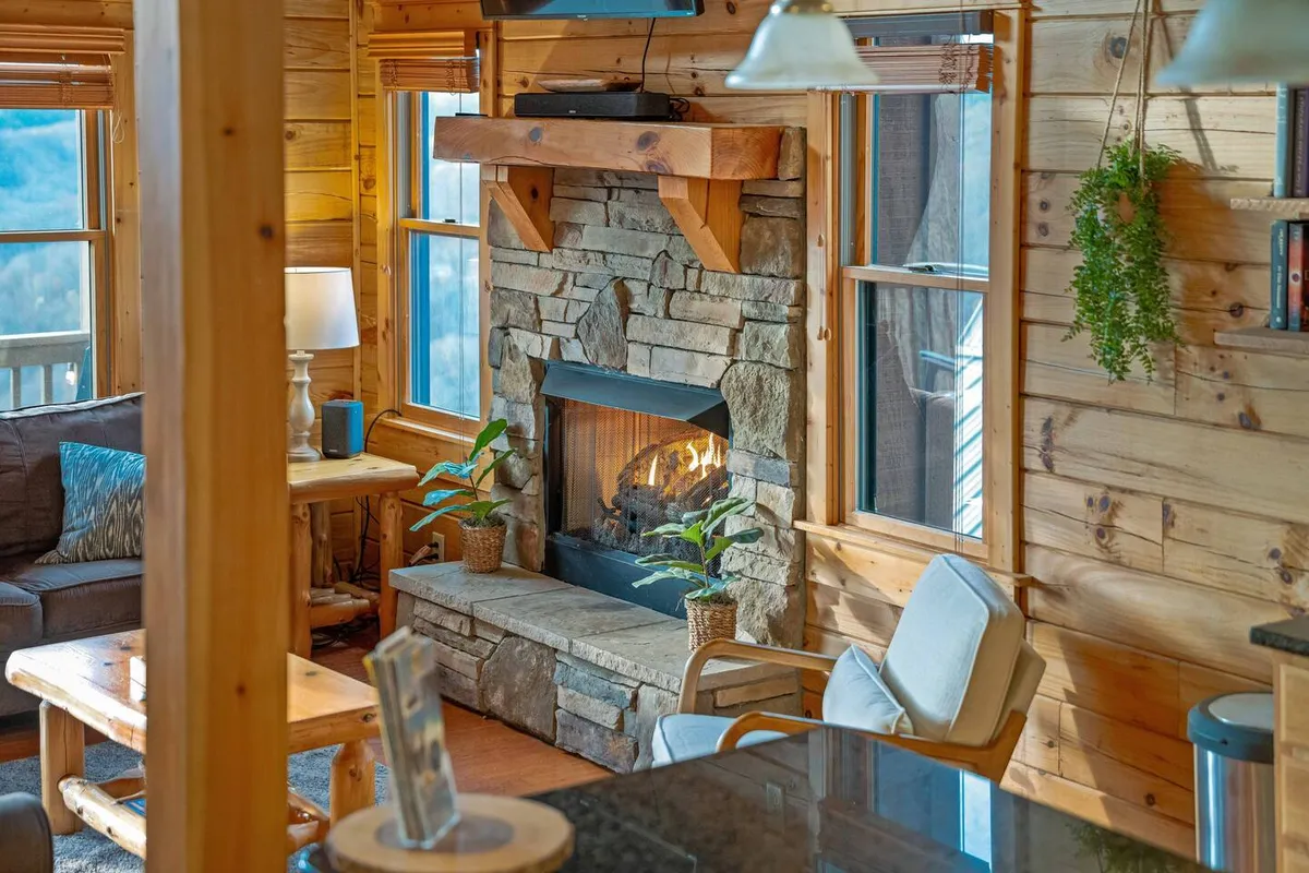 A cozy seating area is centered around a striking stone fireplace, with large windows revealing dramatic mountain vistas. The modern furnishings and warm wood tones create a blend of comfort and style, inviting guests to relax, socialize, or take in the view.