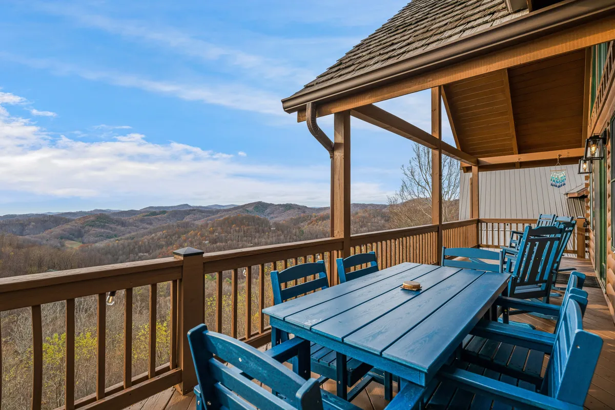 A spacious wooden deck with a blue table and chairs, set against a backdrop of rolling mountains. The setting is perfect for dining outside and relaxing while taking in panoramic mountain views. The wooden railing frames the view, and the atmosphere is tranquil and inviting.
