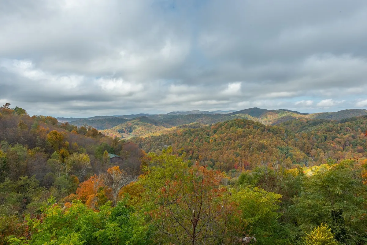 The image displays rolling hills blanketed in autumn foliage beneath a cloudy sky. The expansive landscape offers a sense of the vast wilderness and rich colors visitors encounter while hiking the local trails.