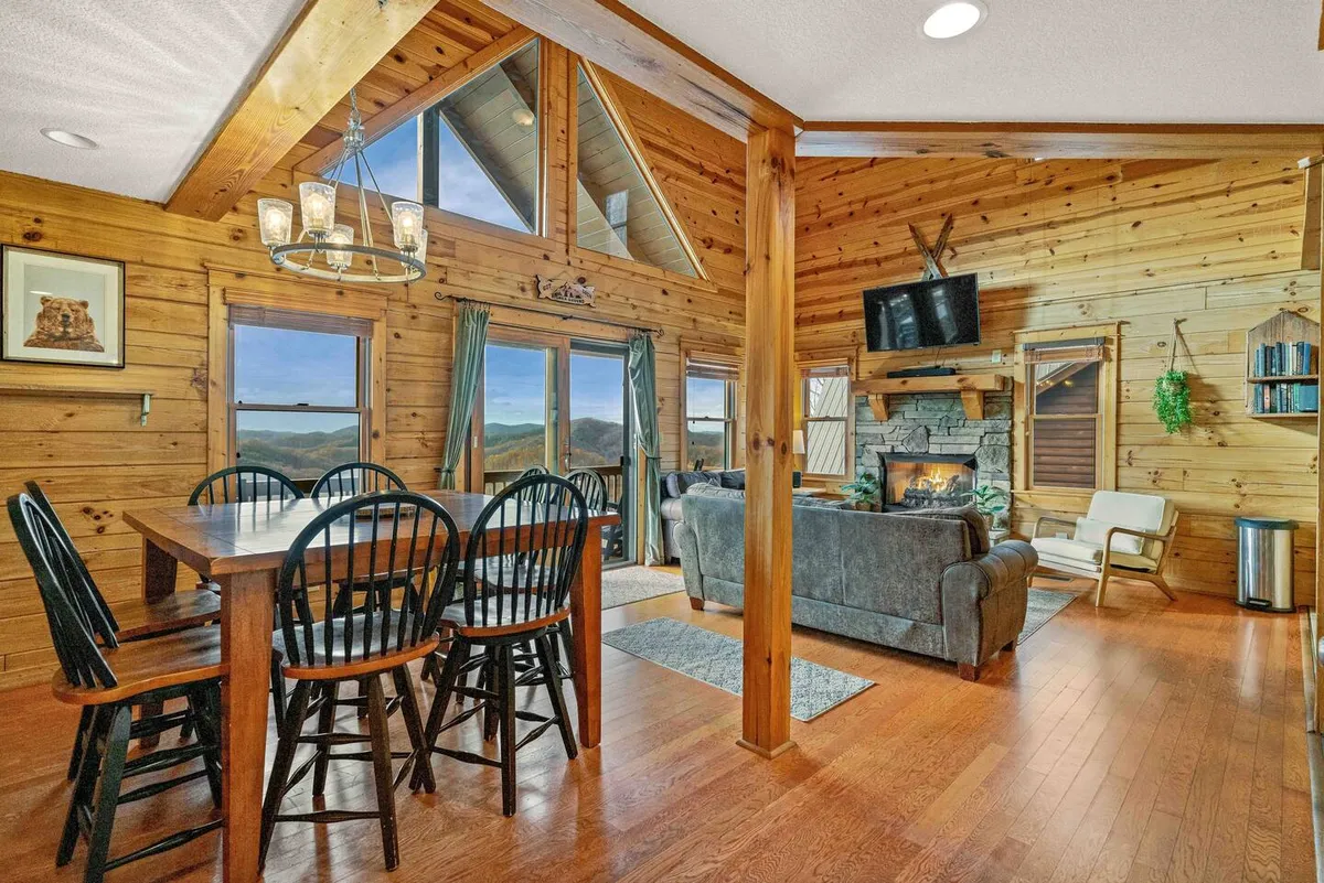 A spacious open-concept living and dining area features a large wooden table set for a meal, a comfortable sofa, and a stone fireplace. Oversized windows frame sweeping mountain views, flooding the room with natural light. The inviting atmosphere is enhanced by contemporary furnishings and cozy accents, perfect for gatherings.