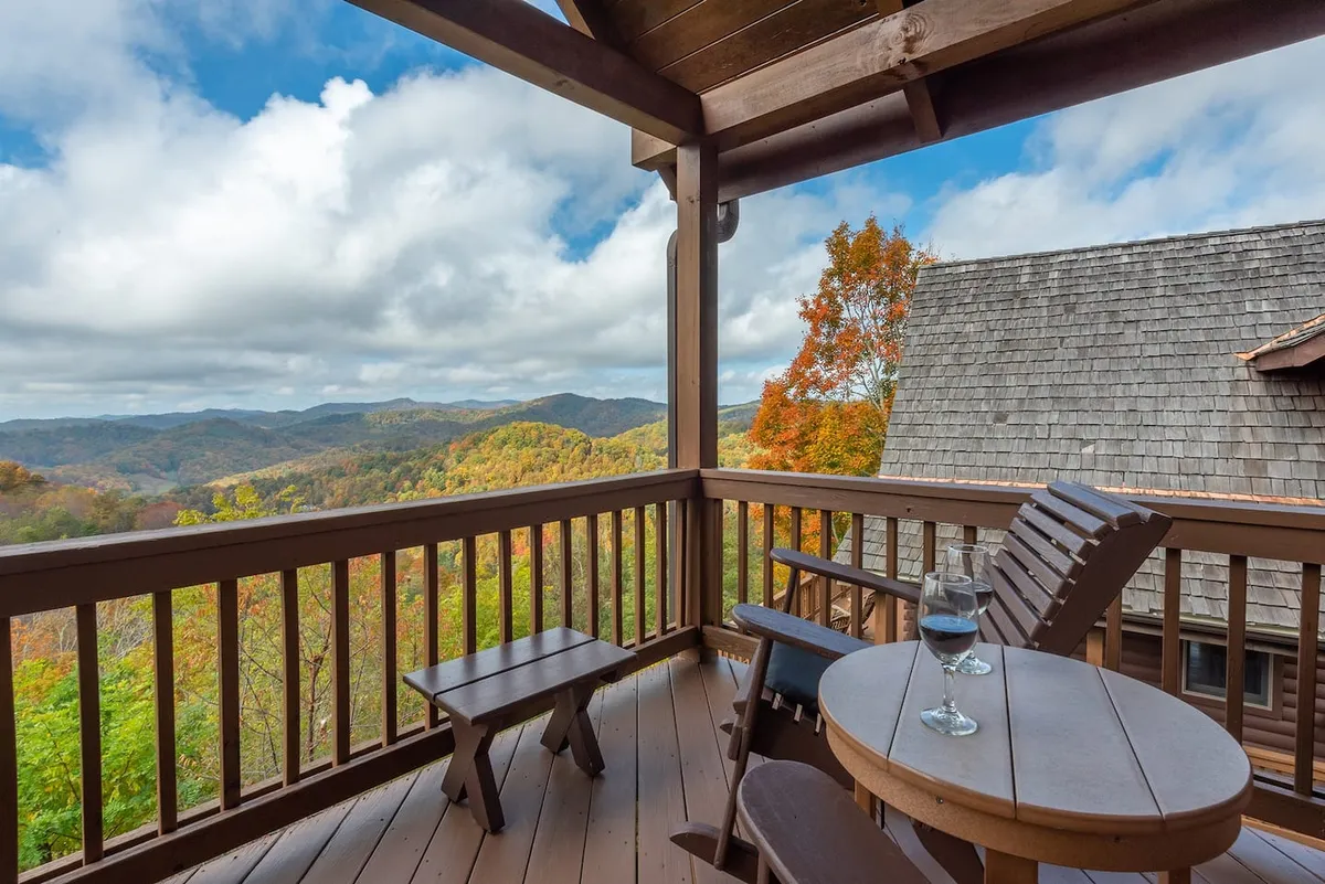 This exterior shot shows a wooden balcony overlooking the expansive Blue Ridge Mountains at sunset, with two wine glasses set on the railing, suggesting relaxation and conviviality. The scene is serene yet festive, with soft golden light draping the landscape and the distant ridges providing a dramatic view.