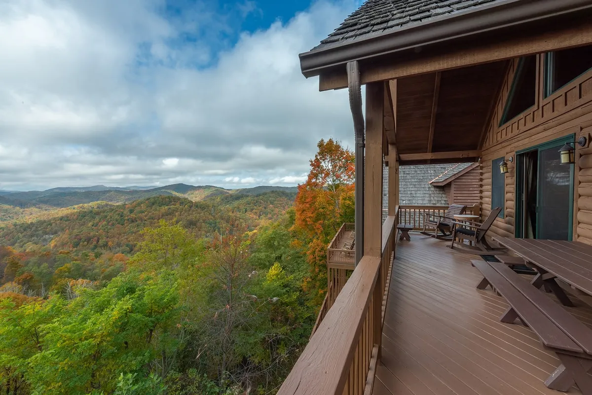 This image features a spacious wooden deck attached to a log cabin, with comfortable seating and a backdrop of autumn foliage and sweeping mountain vistas. The deck is set up for communal gatherings, suggesting both relaxation and celebration in a natural setting.