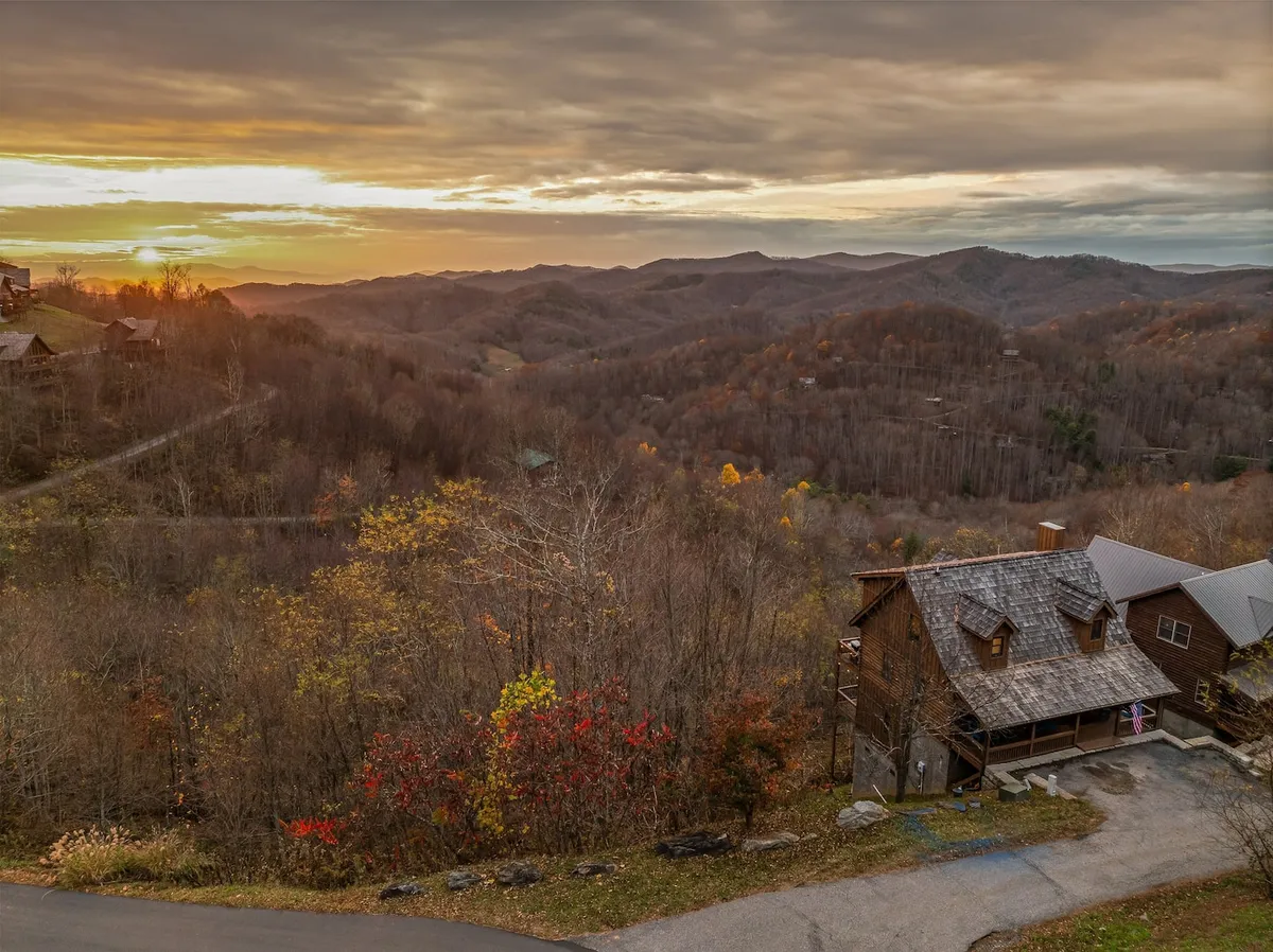 A breathtaking sunset is captured behind mountain peaks, drenching the scene in hues of lavender and gold. Autumn foliage and a wooden cabin appear in the foreground, deepening the sense of peaceful seclusion.