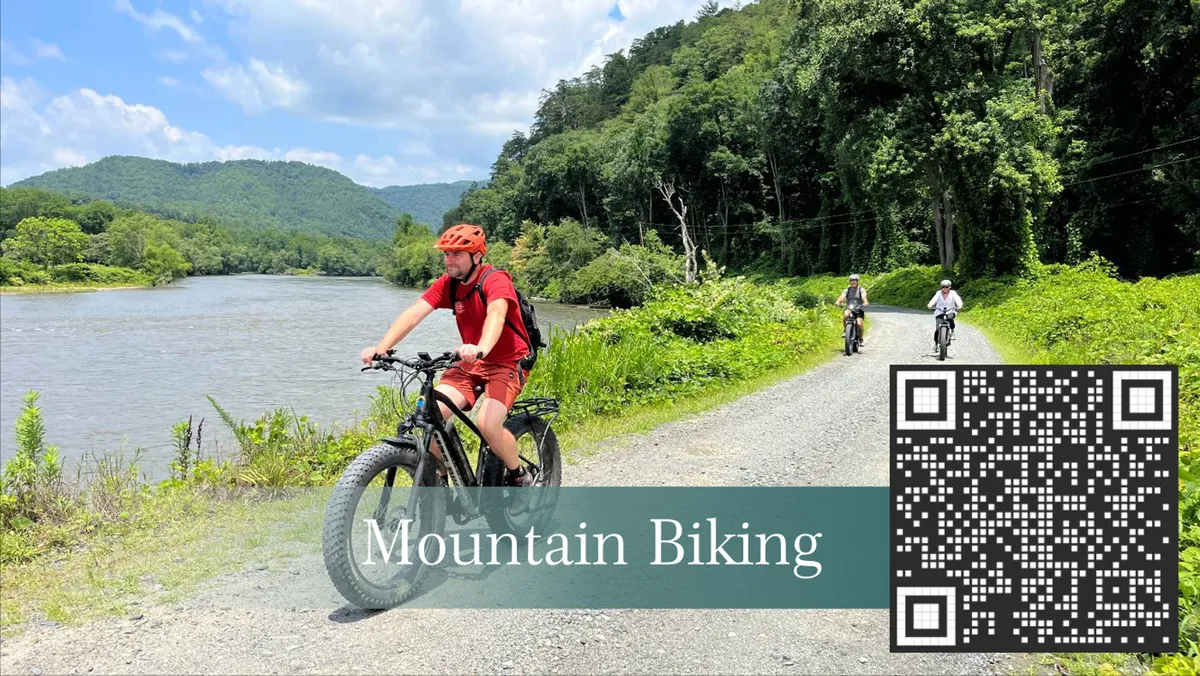A cyclist rides a mountain bike along a riverside trail, framed by lush greenery and a dramatic mountain backdrop. The scene is energetic and showcases active adventure in the Blue Ridge landscape.
