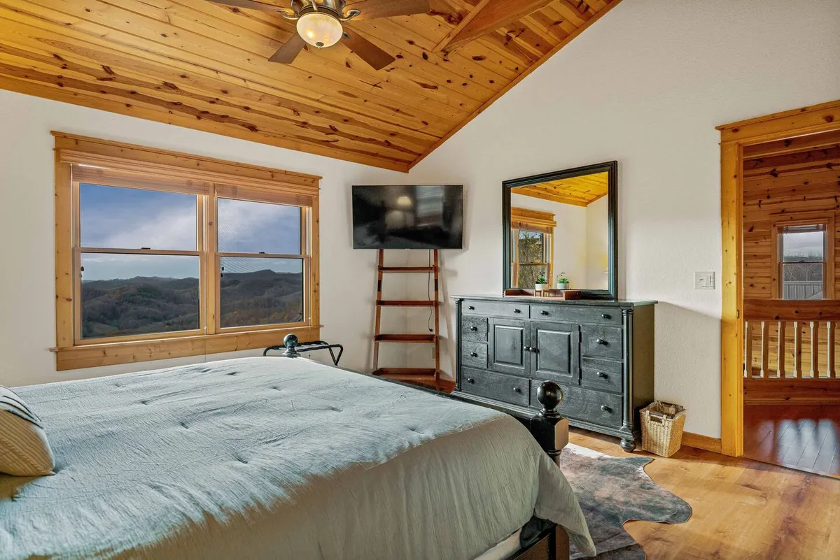 A cozy bedroom featuring a bed with a light blue comforter, large windows providing breathtaking scenic views, and a wooden ceiling. The space feels airy and bright, inviting relaxation while connecting guests to the natural surroundings.