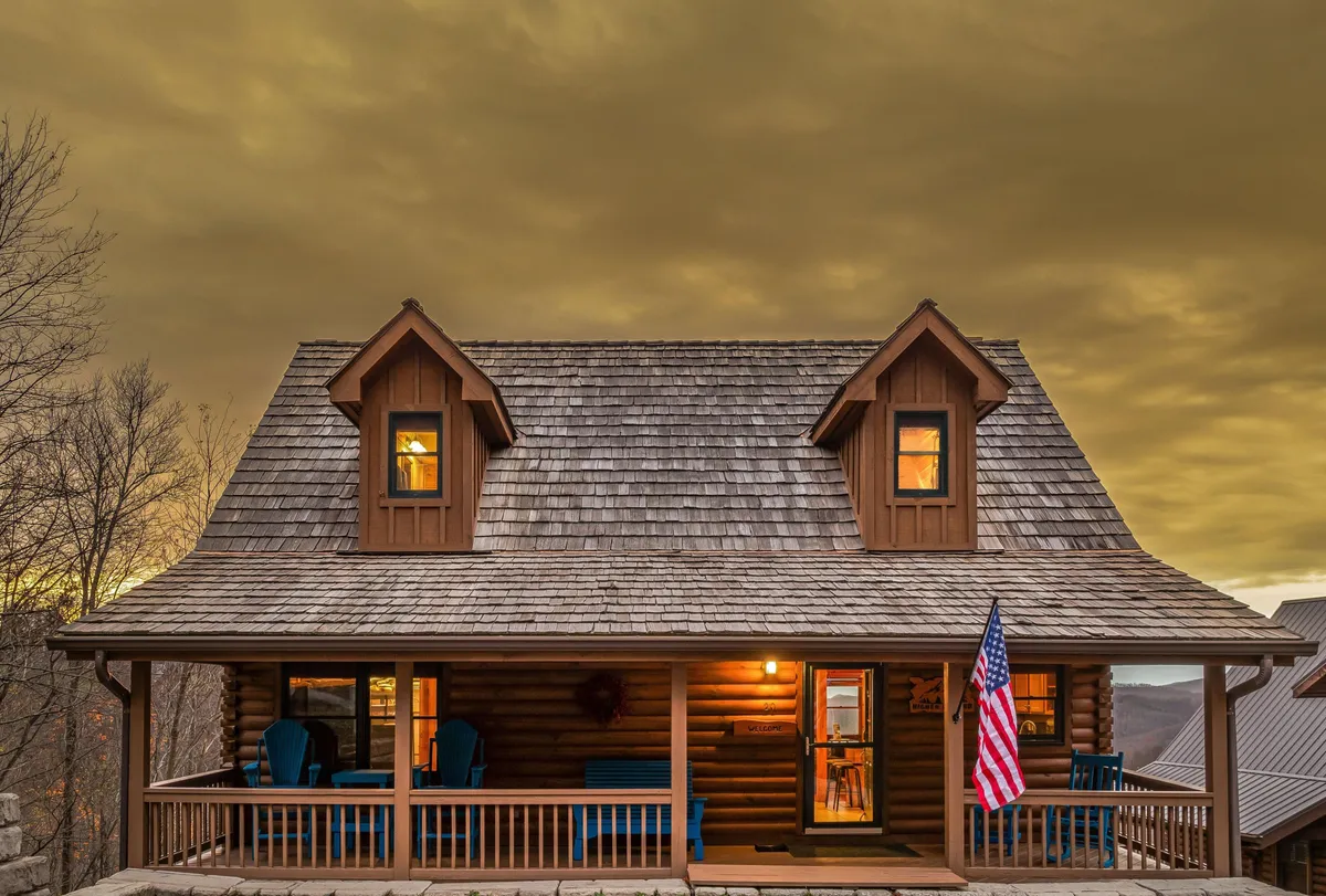 This image features a welcoming log cabin with a front porch, comfortable seating, and the warm glow of lights from the windows. The American flag waves gently, and the setting sun casts a golden hue over the scene, evoking a sense of arrival and togetherness.