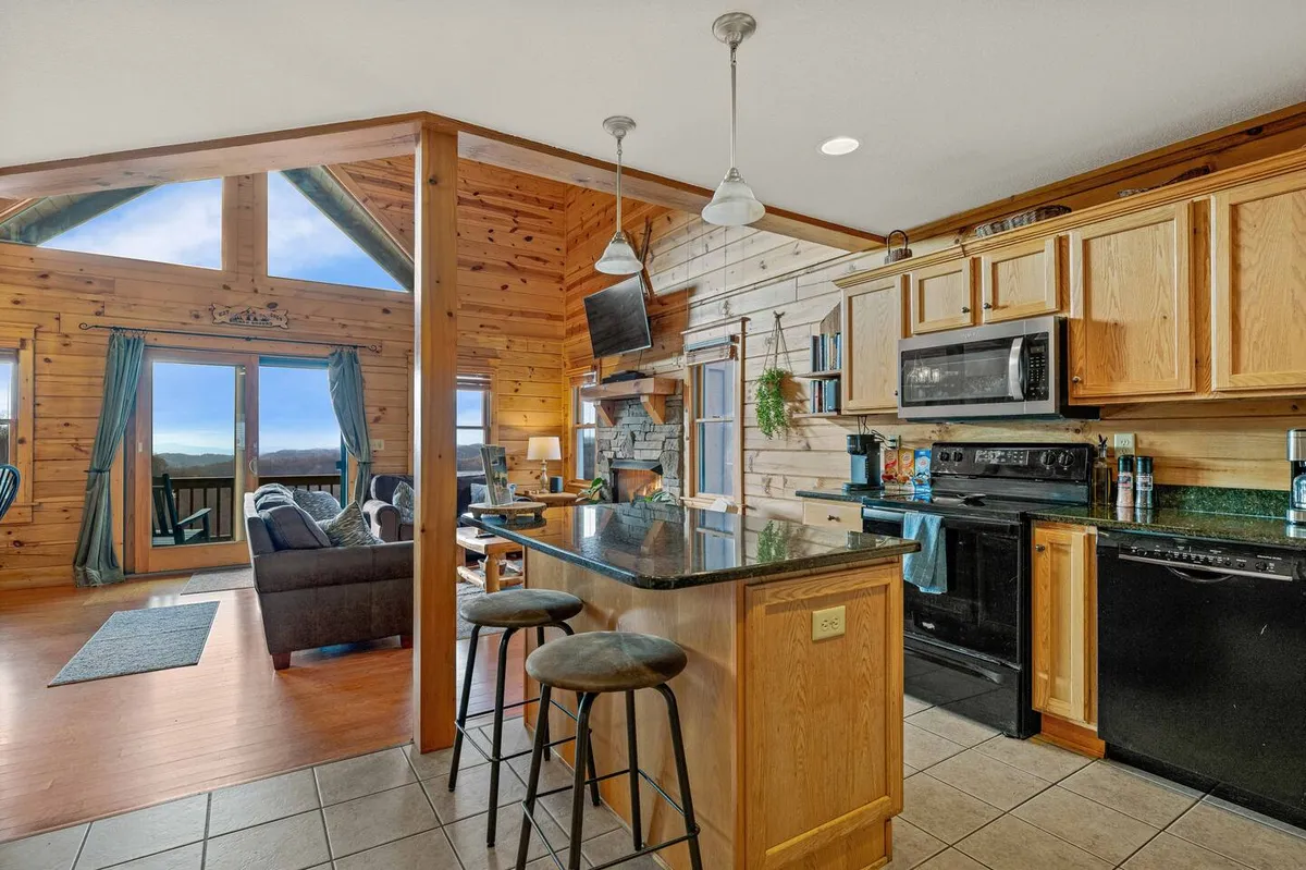 The open living space seamlessly blends a modern kitchen with a large central island, a relaxed seating area, and a stone fireplace. Tall windows provide a stunning mountain backdrop, and the space is furnished with plush, contemporary seating and wooden accents. The layout encourages effortless movement from kitchen to living area.