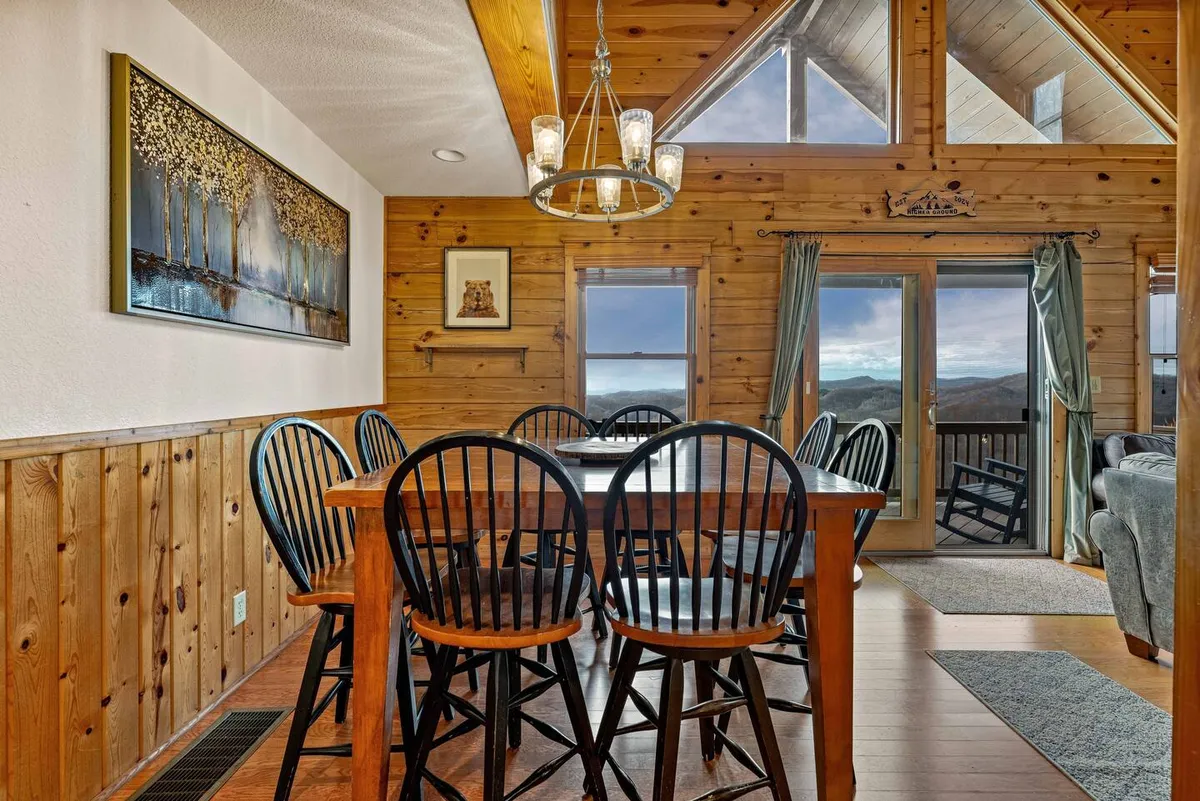 This image captures a warm, wood-paneled dining area with a large table set beneath a chandelier. Expansive windows frame a mountain view, while soft lighting and rustic decor create a cozy, communal atmosphere. The room is clearly designed for gatherings, with ample seating for groups and a sense of welcoming openness.