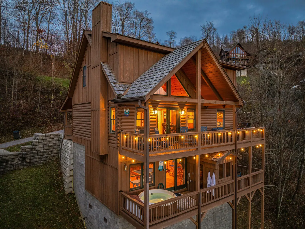 The image showcases the exterior of a modern mountain cabin at dusk, with a spacious wooden deck illuminated by warm string lights. A bubbling hot tub sits invitingly on the deck, framed by sweeping sunset views over the rolling Blue Ridge Mountains. Large windows glow with interior light, hinting at the cozy comfort inside and creating a welcoming, serene atmosphere. The setting evokes relaxation and togetherness, perfect for evening gatherings or stargazing under the open sky.